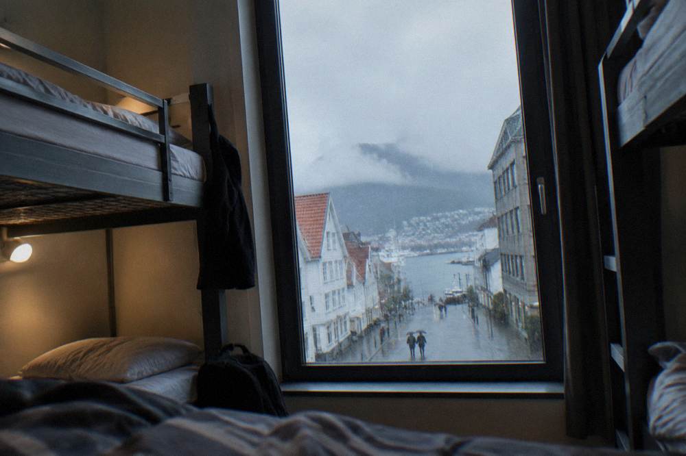 A hostel dormitory room with bunk beds and a window showing a rainy Norwegian city street