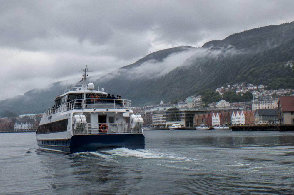 A tour boat departing from the inner harbour in Bergen with mountain ridges behind the city