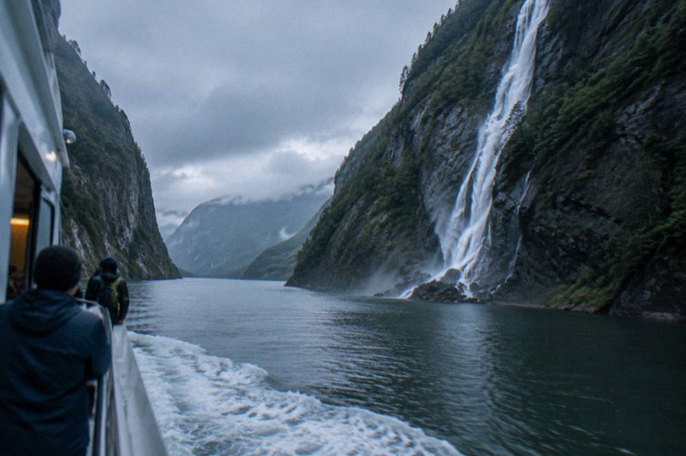 A sightseeing boat passing through the narrow Mostraumen strait between steep rock walls with a waterfall pouring down one side