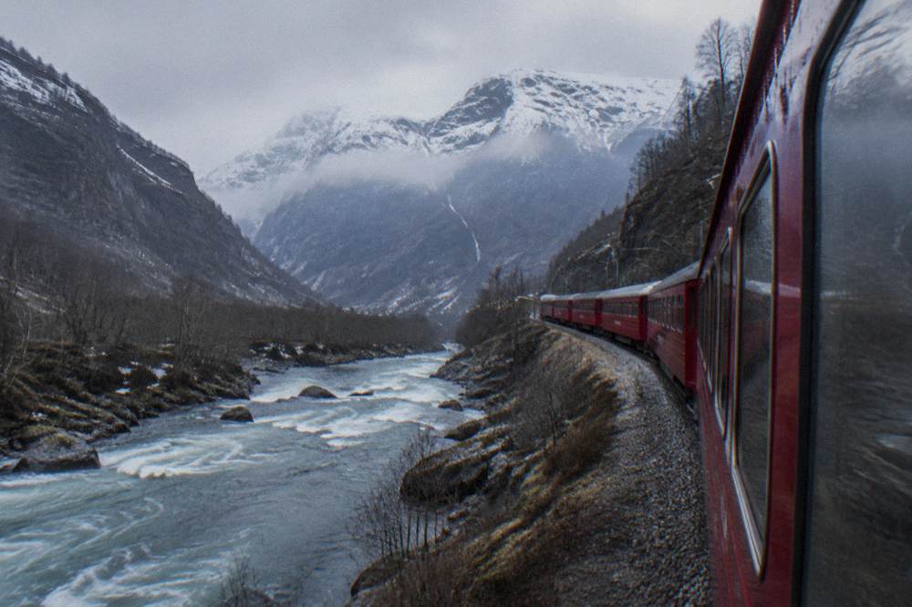 A red railway coach descending the steep Flåm valley beside a fast river with snow-capped mountains above