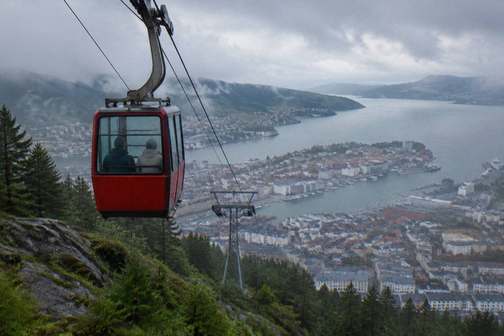 Two passengers in a red cable car cabin ascending above Bergen with the city and fjord spreading out below
