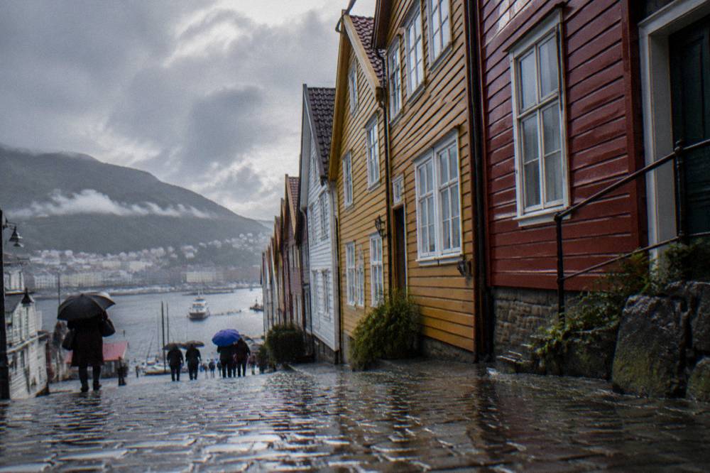 Wet cobblestone street in central Bergen with people under umbrellas and water beading on colourful building fronts