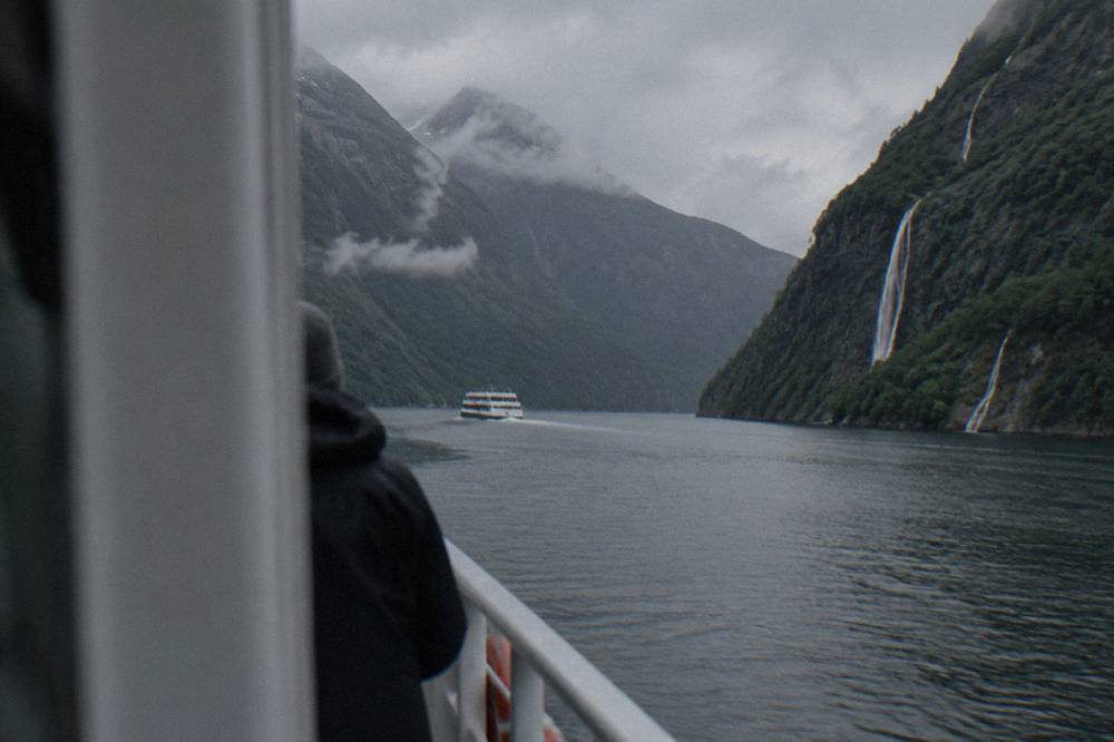 Small cruise boat on a narrow fjord arm between steep forested cliffs with waterfalls