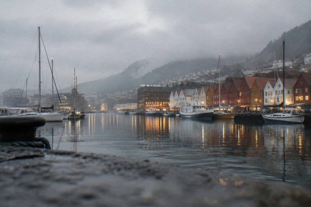 Evening view across Bergen harbour with moored boats, lit warehouses and reflections on calm water