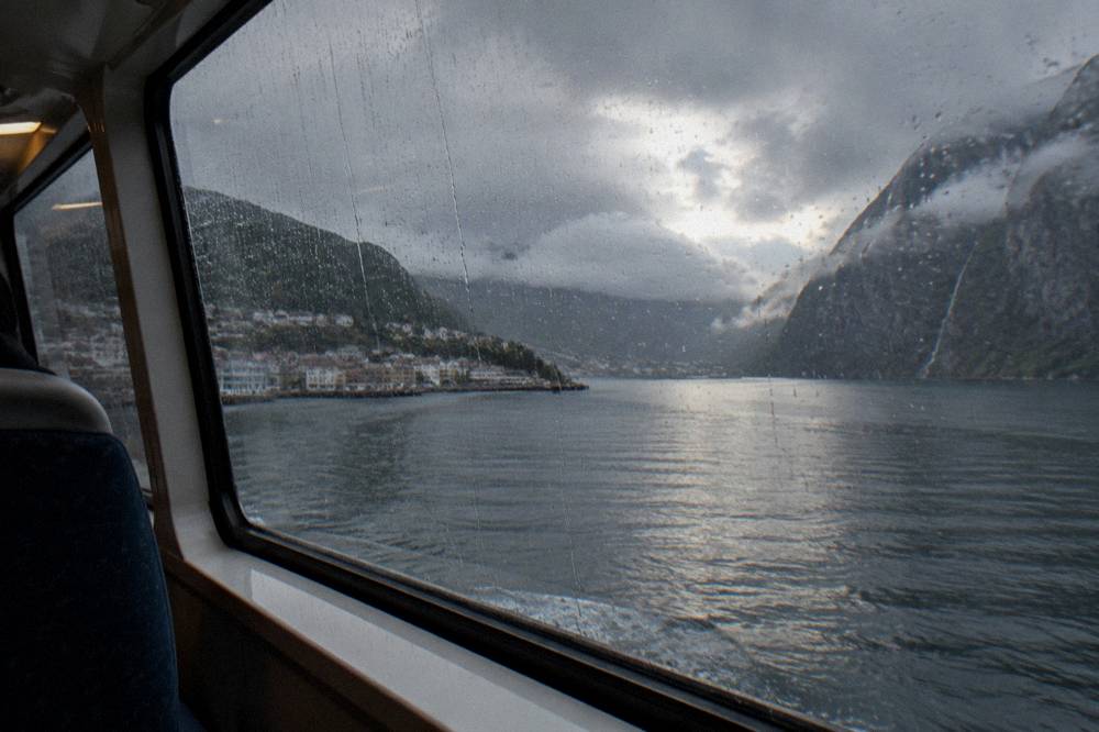 Rain streaking a boat cabin window with dark fjord water and cliffs outside
