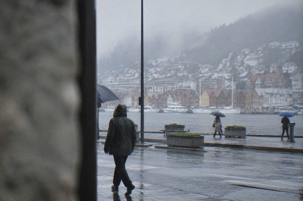 A walker in a rain jacket crossing a wet street in central Bergen with umbrellas nearby