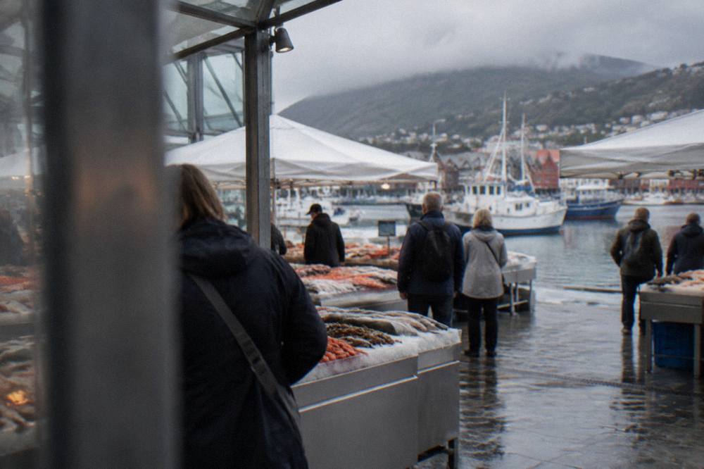 Tourists browsing stalls under white tents at the Bergen Fish Market, with the harbour and moored boats behind them.
