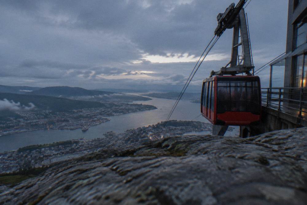 The red Ulriken cable car arriving at the upper station on Mount Ulriken, with Bergen and the fjord spread out below under broken cloud.