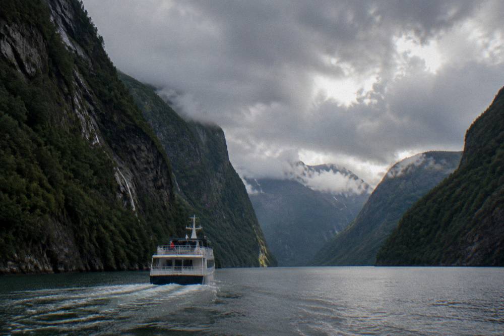 A small cruise boat heading up a narrow Norwegian fjord channel with steep forested cliffs on both sides.