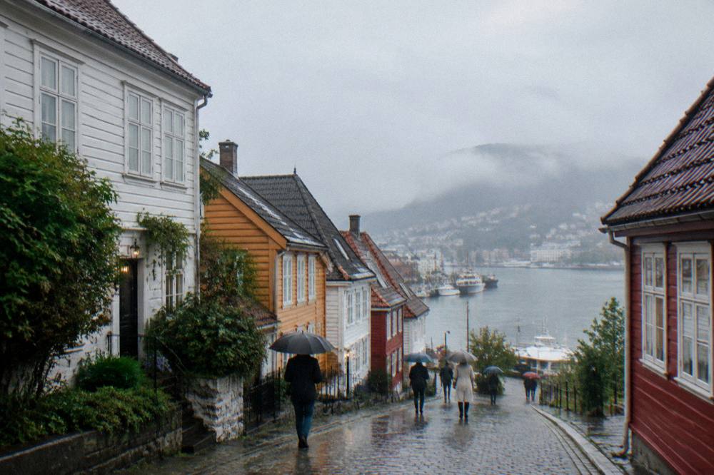 A rain-slicked cobblestone street between colourful wooden houses in central Bergen, with people under umbrellas.