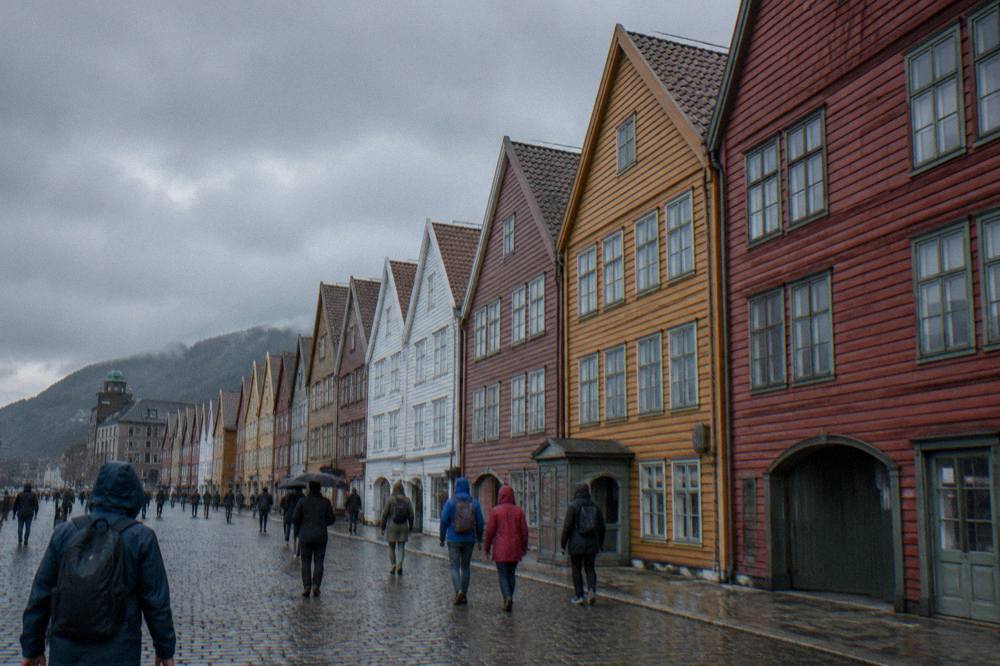 Visitors with rain jackets walking between the coloured wooden buildings of Bryggen in Bergen under overcast skies.