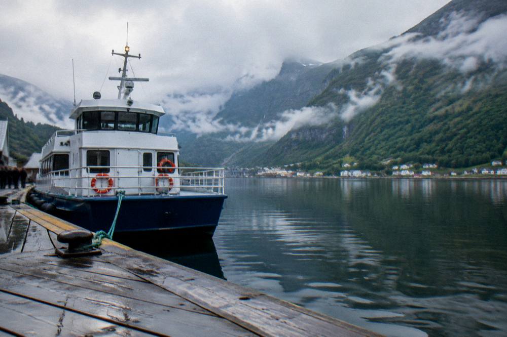 A passenger boat moored at a wooden jetty on a calm Norwegian fjord, with steep green mountainsides reflected in the still water.